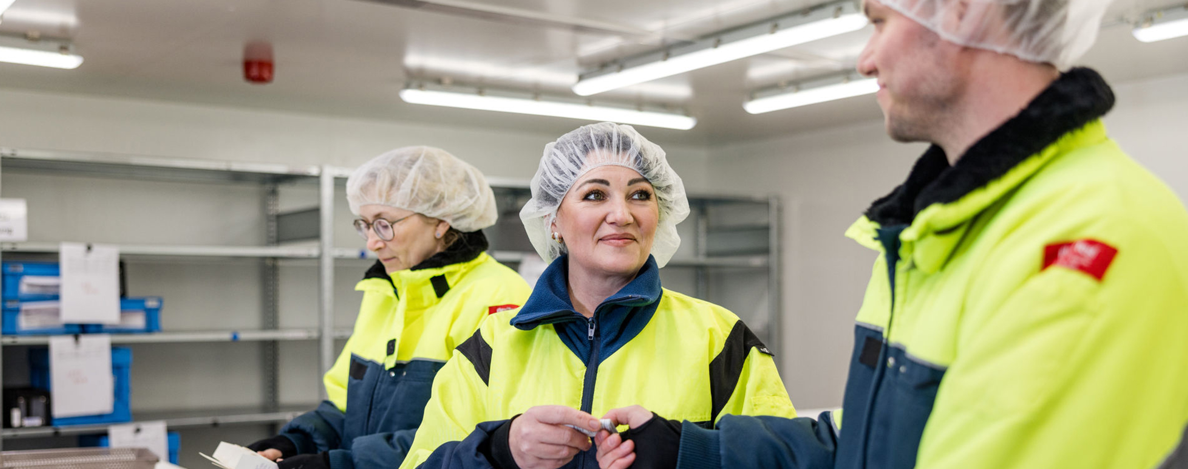 Es sind drei Personen, zwei Frauen und ein Mann, in leuchtend gelben Sicherheitsjacken und Haarnetzen in einem Kühlbereich zu sehen. Der Fokus liegt auf der Frau und dem Mann im Vordergrund, die sich unterhalten und sich ein Papier übergeben. Die Frau im Hintergrund schaut auf eine Medikamentenverpackung.