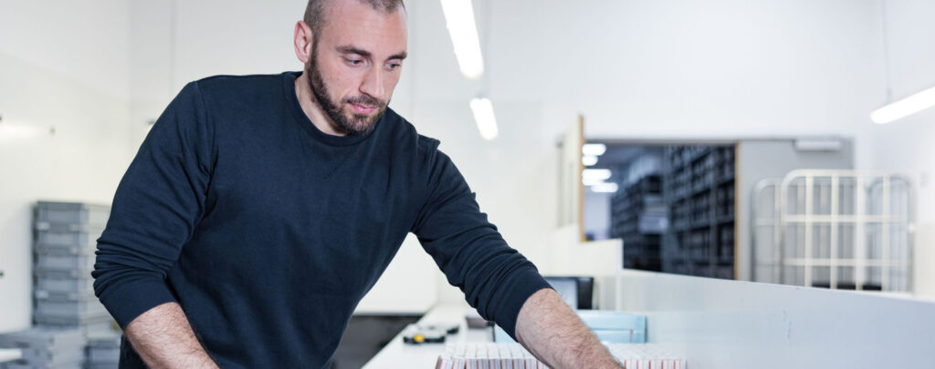 Ein Mann arbeitet an einem Arbeitsplatz in einer lagerartigen Umgebung. Er steht gebeugt über einen Tisch, auf dem Utensilien, wie Medikamentenverpackungen zu sehen sind.