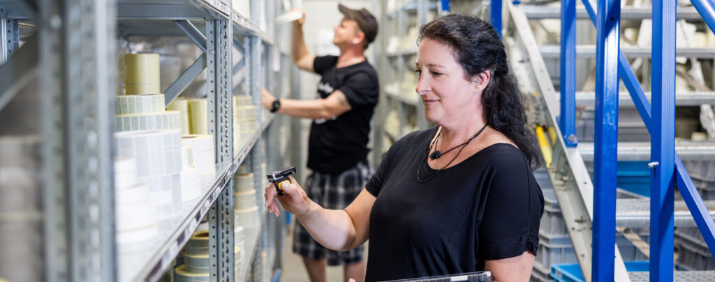 Zwei Personen, ein Mann und eine Frau stehen vor Regalen in einer Lagerumgebung, wobei die Frau im Vordergrund ein Handscanner-Gerät verwendet. Der Mann im Hintergrund hält Produkte aus dem Regal in der Hand.